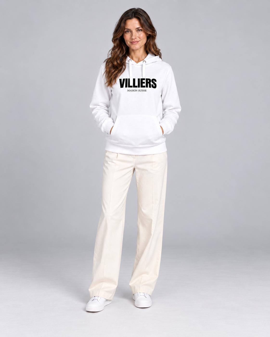 Frau trägt weissen Premium Hoodie von Villiers Maison Suisse zu cremefarbener Hose, Ganzkörper von vorne
